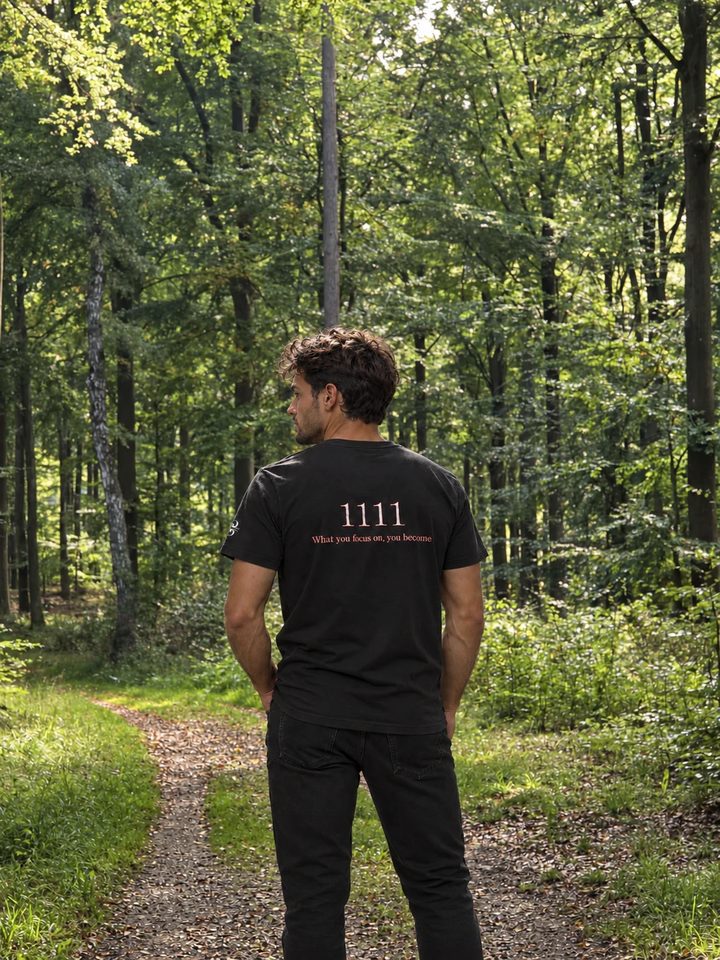 En mand i skoven set bagfra i en sort t-shirt med 1111 og teksten What you focus on, you become på ryggen.